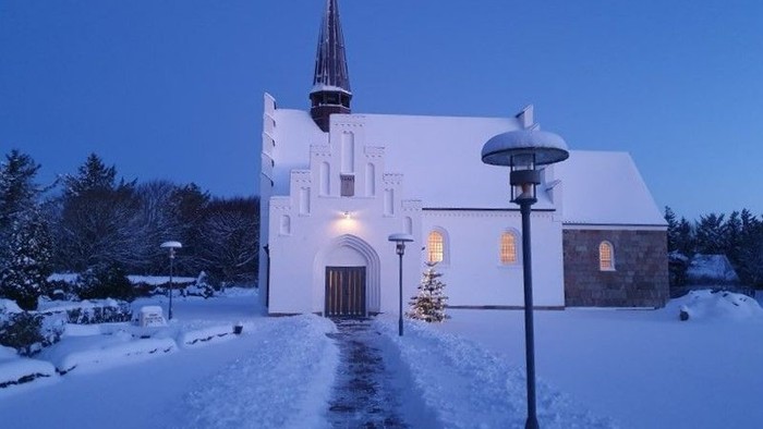 En snede kirke med lysende vinduer og tårn i snefaldende landskab