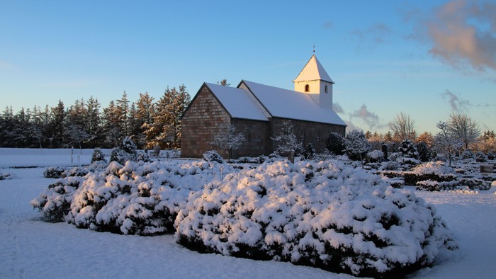 En snedækket kirke med tårn og en lille bygning ved siden af, omgivet af snebergede buske og træer under et klart blå himmel.