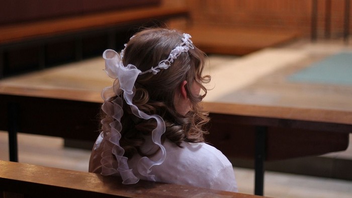 Ein junges Mädchen in einem weißen Kleid mit einem verzierten Kopfschmuck sitzt während einer Zeremonie in einer Kirchenbank.
