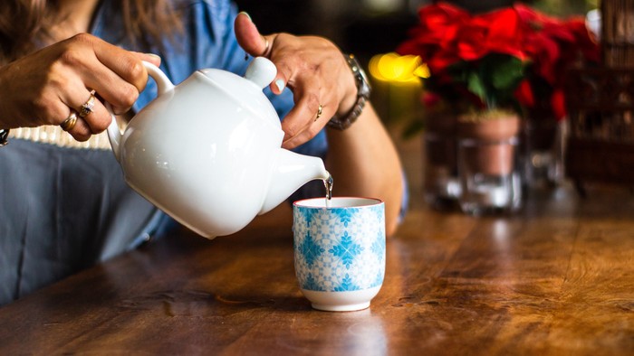 Eine Person gießt Tee aus einer weißen Kanne in eine blaue Tasse auf einem Holztisch.