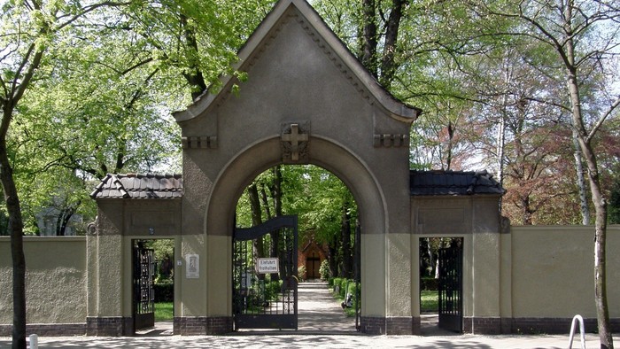 Das Bild zeigt einen Torbogen aus Stein mit einem Kreuz darüber, umgeben von Bäumen und einem Zaun.