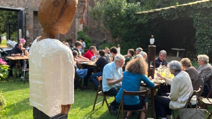 Holzskulptur auf Rasenfläche, Menschen sitzen an Tischen, Ziegelgebäude im Hintergrund