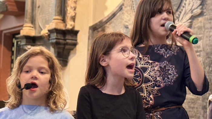 Drei Personen, die gemeinsam auf der Bühne mit Mikrofonen singen, darunter zwei Kinder und ein Erwachsener.
