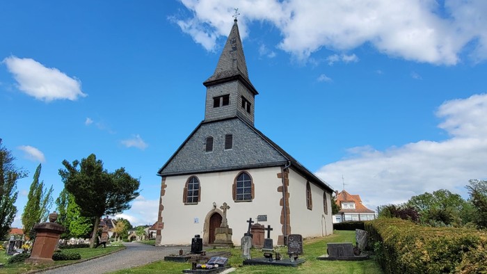 Idyllische Kirche mit hohem Turm, umgeben von einem ruhigen, gepflegten Friedhof unter blauem Himmel.