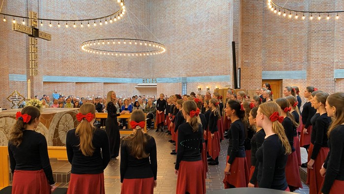 En gruppe unge piger i koordineret sort og rød påklædning, der synger i en rummelig, veloplyst kirke.