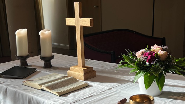 Eine ruhige Altar-Anordnung mit Kerzen, einem Holzkreuz, einem offenen Buch, Blumen und einer Schale mit Räucherwerk.