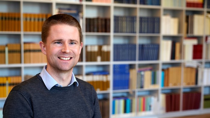 En smilende mand står foran et velassorteret, farverigt bibliotek med bogreoler.