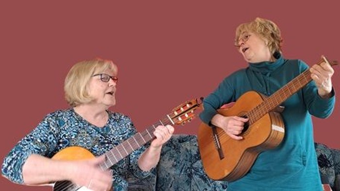 Zwei Frauen spielen fröhlich gemeinsam Ukulele in passenden Outfits vor einem lebhaften Hintergrund.