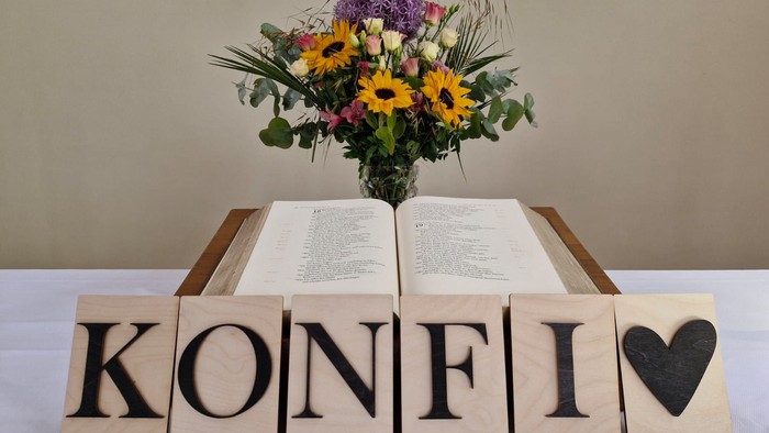 Holzbausteine bilden das Wort „KONFI“ mit einem Herz, platziert unter einem offenen Buch und einer Blumenanordnung.