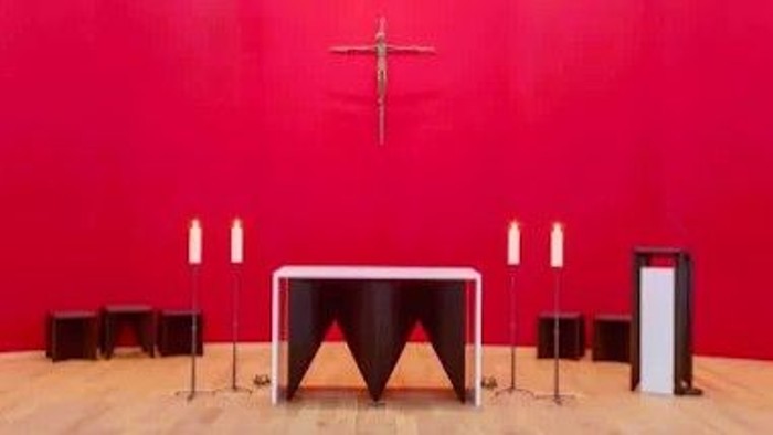 Moderner Altar mit Kruzifix vor einer roten Wand und brennenden Kerzen.