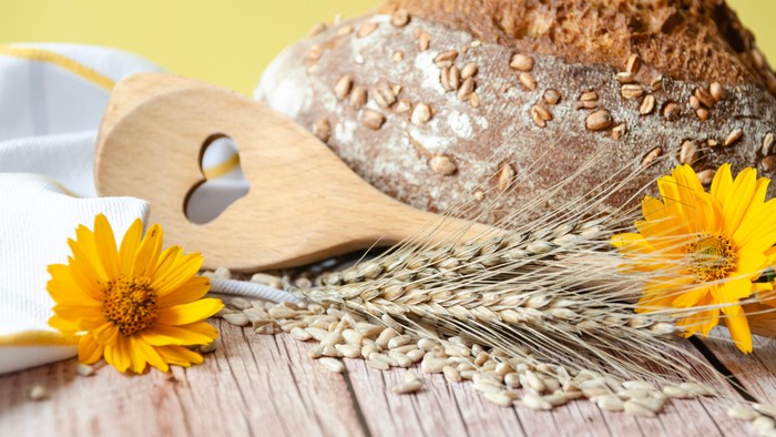 Das Bild zeigt ein Holzlöffelchen neben Brot, Getreide und gelben Blumen.