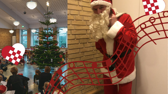 Santa Klaus står ved en mur med juledekorationer og en juletree.