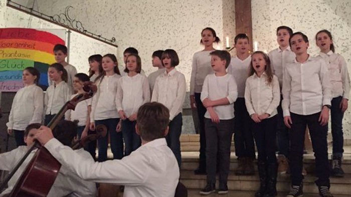 Ein Jugendchor singt, begleitet von einem Cellisten, in einer kirchenähnlichen Umgebung.