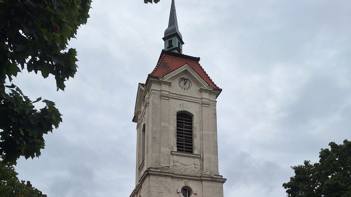 Kirche mit hohem Kirchturm und Kreuz, umgeben von Bäumen und Gras.