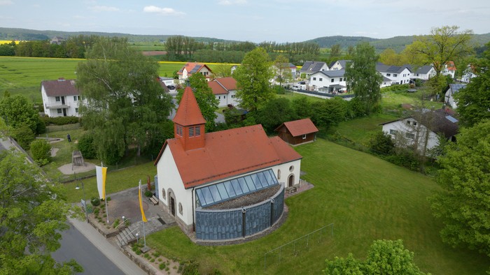 Ein weißes Gebäude mit rotem Dach in ländlicher Umgebung, umgeben von Grünflächen und weiteren Häusern.
