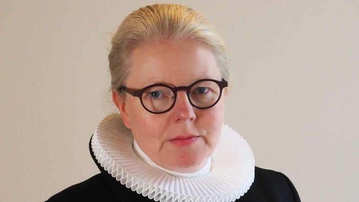 En kvinde med blond hår og briller bærer en sort jakke med hvid krage.