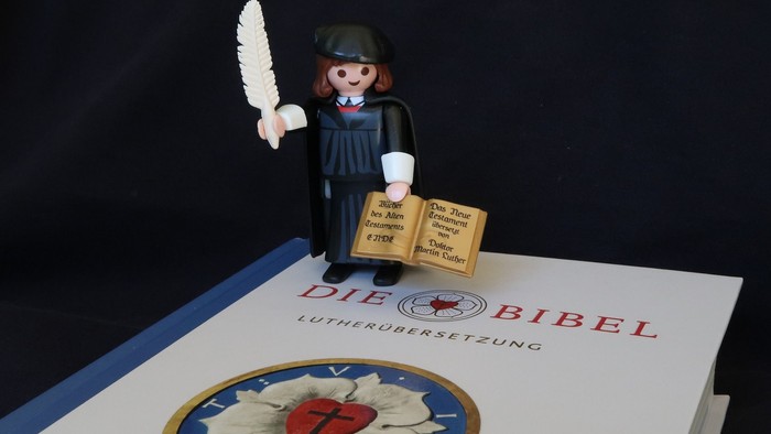 Eine Playmobil-Figur hält eine Feder und ein Buch über der Die Bibel, die auf einem Tisch liegt.
