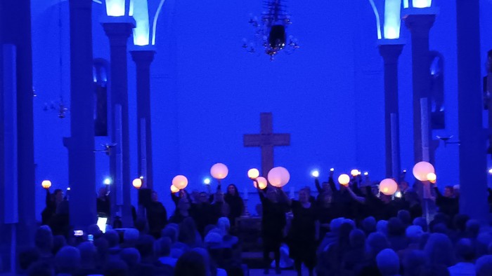 En skare holder glødende lanterner under en højtidelig procession i en svagt oplyst, blåtonet kirke.