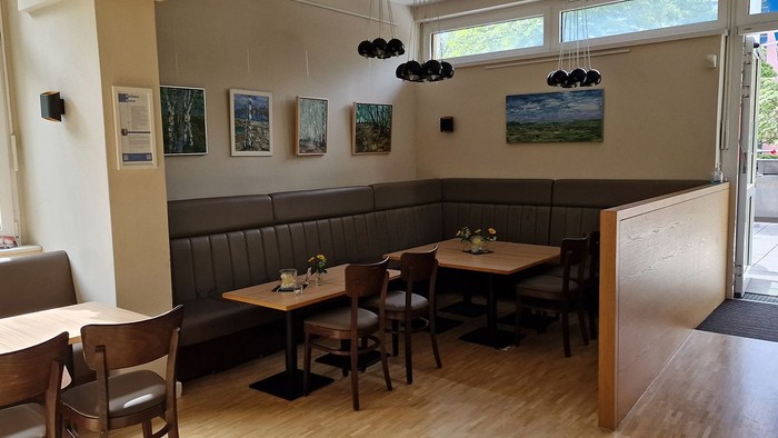 Gemütliches Café mit Holzboden, Tischen und Stühlen, Kunst an den Wänden und großen Fenstern