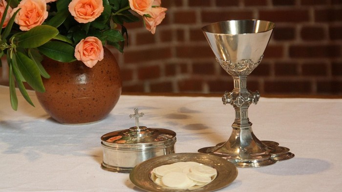 Kelch und Hostienschale auf einem Altar. Im Hintergrund eine Vase mit rosafarbenen Rosen.