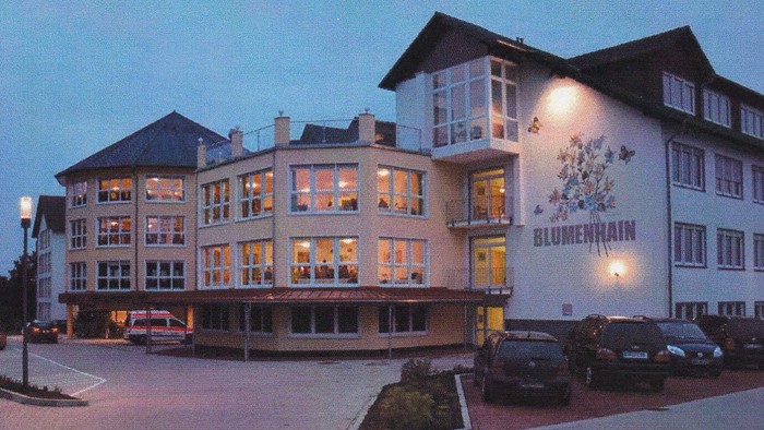 Modernes Gebäude namens Blumenhain mit beleuchteten Fenstern, Autos davor geparkt bei Dämmerung.