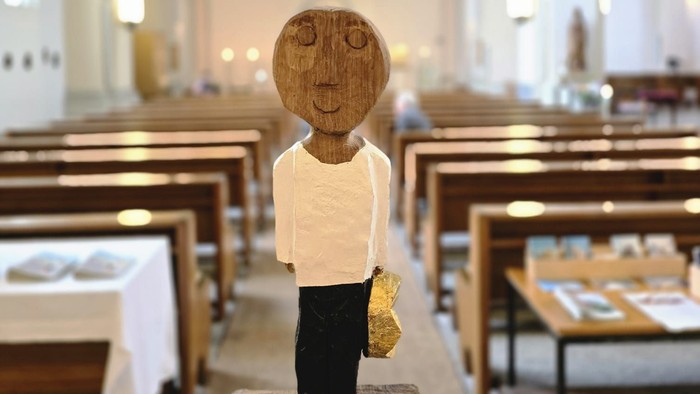 Holzfigurine im Kirchenschiff, die einen kleinen Gegenstand hält, auf einem Steinsockel platziert.