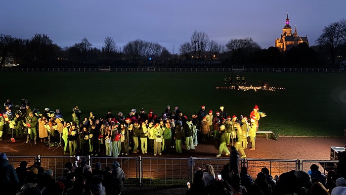 Eine lebhafte Gruppe in koordinierten gelben Outfits tritt bei Dämmerung auf einem Stadionfeld auf, mit einer Burg im Hintergrund.