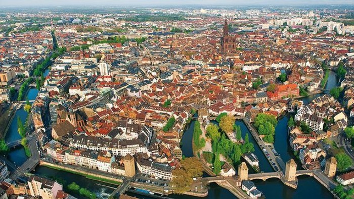 Vue aérienne d'une ville européenne historique avec des rivières sinueuses, des bâtiments aux toits rouges et une cathédrale imposante.