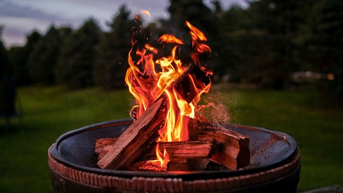 Das Bild zeigt eine Feuerschale auf einer Wiese mit Bäumen im Hintergrund.