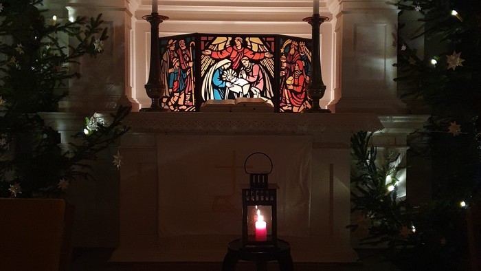 Ein ruhiges Kircheninneres zeigt Buntglasfenster und eine von funkelnden Lichtern und Grünpflanzen umgebene Laterne mit Kerzenlicht.