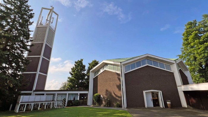 Moderne Kirchenanlage mit hohem Glockenturm in einer üppigen, grünen Außenanlage.