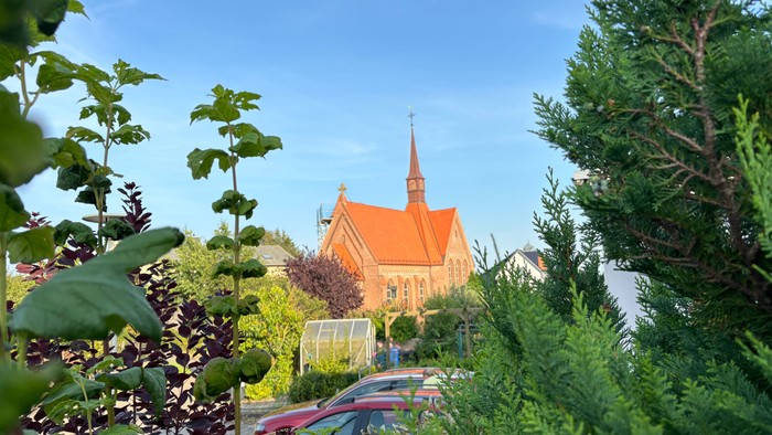 Ein rotes Kirche mit spitzem Turm, umgeben von grünem Gebüsch und parkenden Autos.
