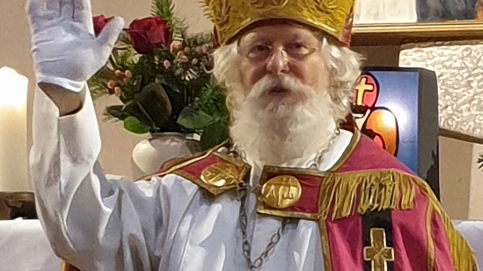 Ein Mann in kirchlicher Kleidung mit goldenem Hut und weißem Bart hält eine Blume hoch
