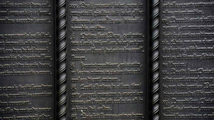 Aufwendig eingravierter Text auf einer dunklen, verdrehten Metalloberfläche, der an alte Schriftzeichen oder Code erinnert.