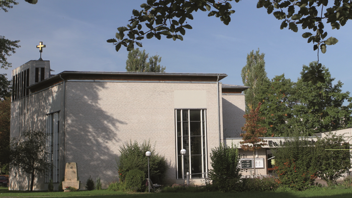 Moderne Kirche mit Kreuz auf dem Dach, umgeben von Bäumen und Grünflächen.