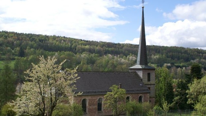 Kirche mit hohem Kirchturm, umgeben von Bäumen und Hügeln.