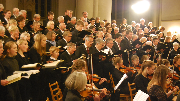 Ein großes Orchester und ein Chor treten gemeinsam in einem prächtigen, hell erleuchteten Konzertsaal auf.