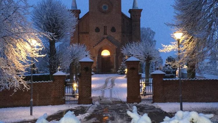 Sneklædt kirke med oplyste vinduer og spir, indrammet af vintertunge træer og buske.