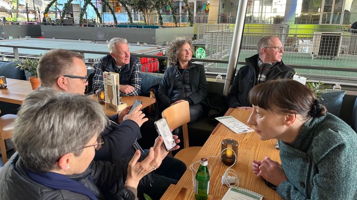 En gruppe mennesker, der socialiserer indendørs på en café, bruger telefoner og nyder drinks sammen.