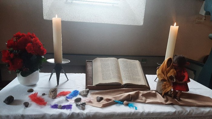Eine ruhige Altargestaltung mit Kerzen, Blumen, einem offenen Buch und verstreuten bunten Federn auf einem bedeckten Tisch.