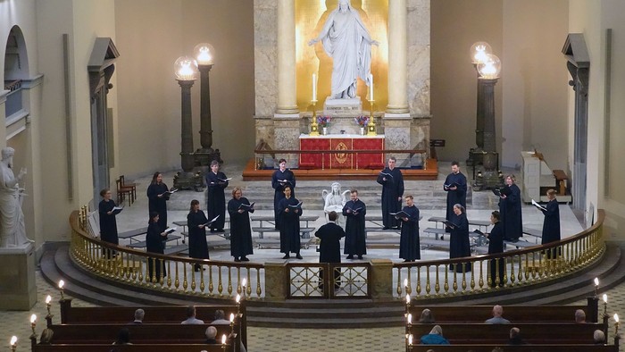 Et kor optræder i en stor kirke under en formel gudstjeneste med alteret som baggrund.