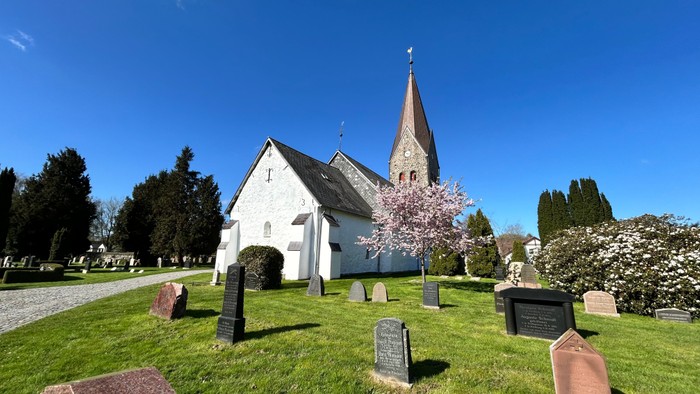 Hyggelig hvid kirke med højt spir omgivet af en rolig, velplejet kirkegård under en klar blå himmel.