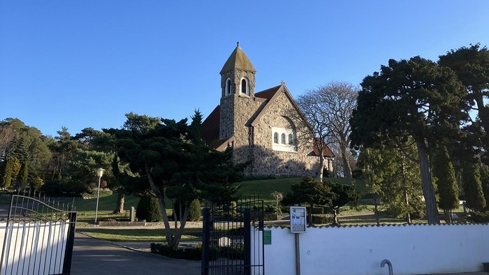 Hyggelig stenkirke med klokketårn omgivet af frodig grøn natur under klar blå himmel.