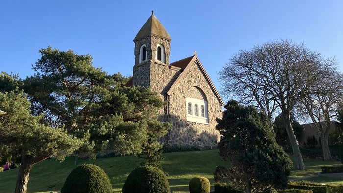 Hyggelig stenkirke med klokketårn omgivet af veltrimmet grønne områder under en klar blå himmel.