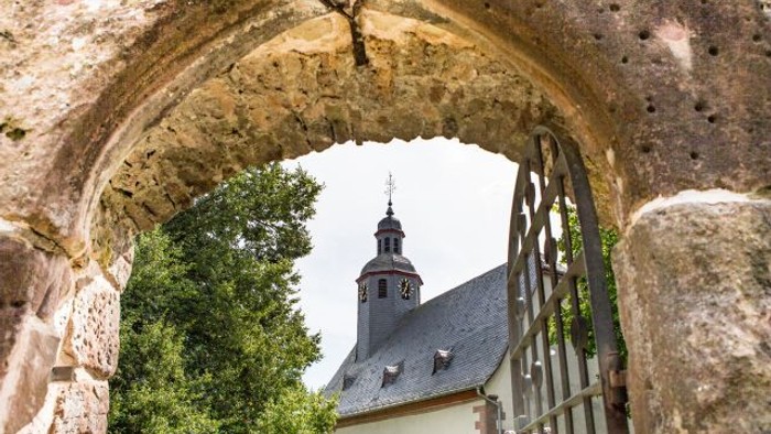 Ein steinerner Torbogen rahlt einen Uhrturm und ein Gebäude mit steilem Dach ein.