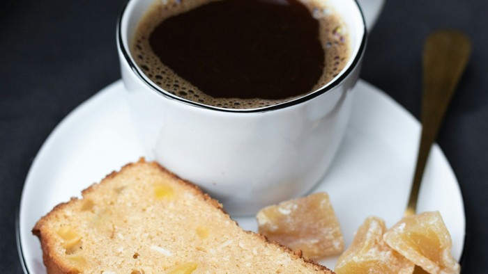 Eine dampfende Tasse Kaffee steht neben einem Stück Zitronen-Gugelhupf und kandierten Früchten auf einem Unterteller.