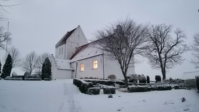 En snedækket kirke med et lysende vindue og nakke træer.