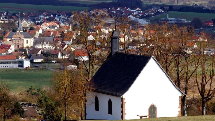Idyllische weiße Kapelle, eingebettet in ein malerisches Dorf auf dem Land, umgeben von sanften Hügeln und kahlen Bäumen.