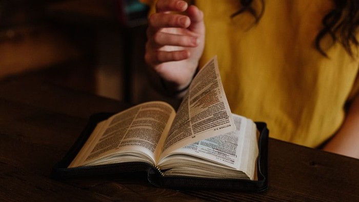 Eine Person liest ein aufgeschlagenes Buch mit gefalteten Händen über den Seiten auf einem Holztisch.