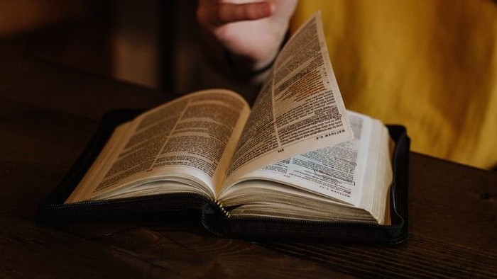 Eine Person liest ein aufgeschlagenes Buch mit gefalteten Händen über den Seiten auf einem Holztisch.